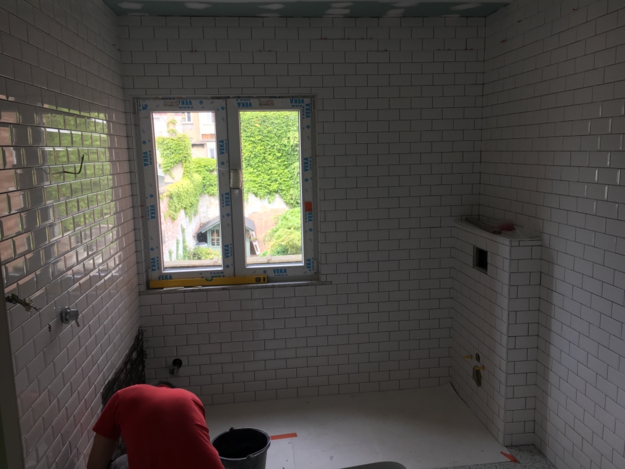 Biała łazienka w trakcie remontu: glazurnik w czerwonej koszulce układa białe, prostokątne płytki na ścianach, widoczne okno z widokiem na zieleń, poziomica na parapecie, wiadro i narzędzia...