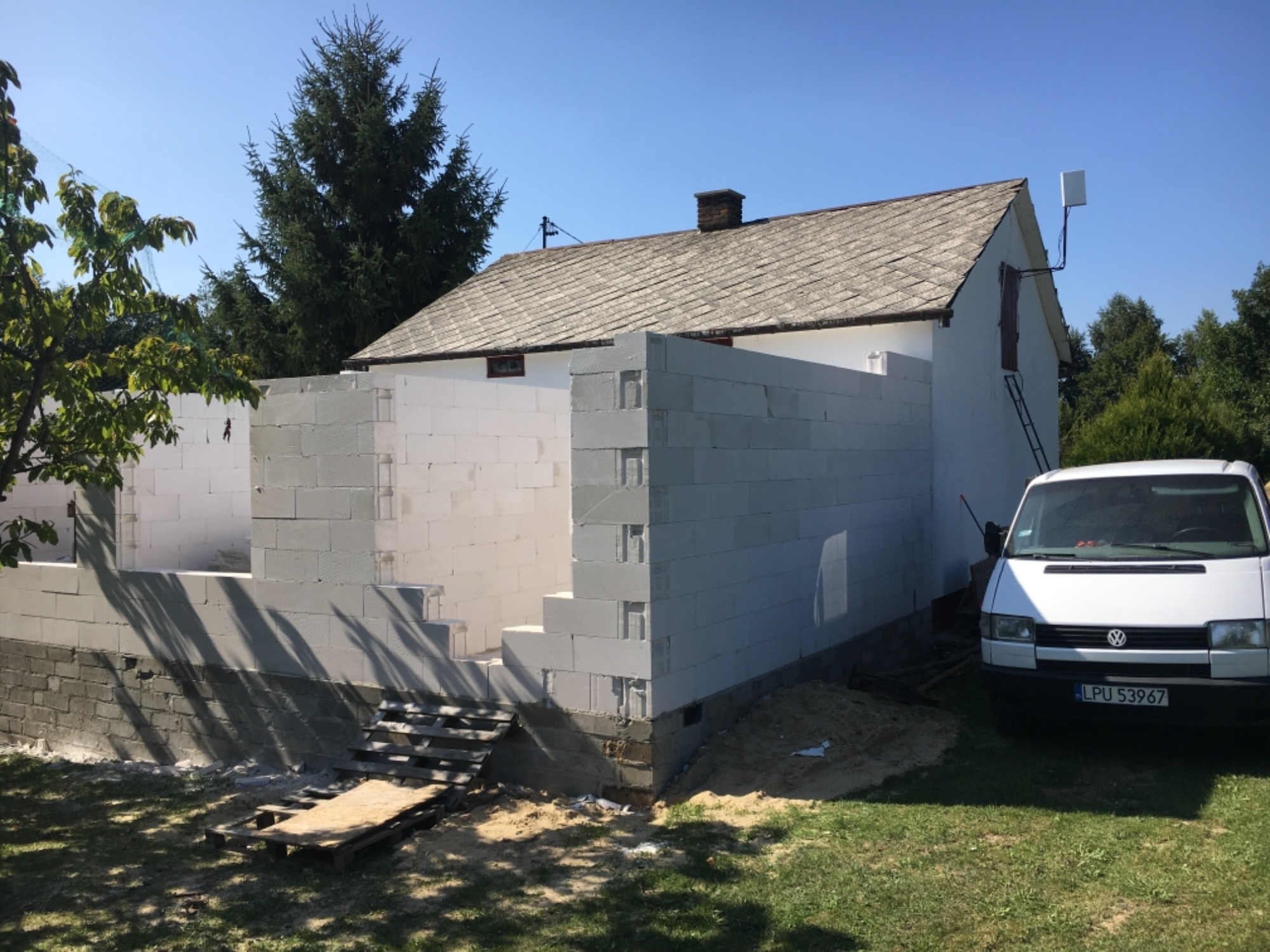 Wznoszenie ścian zewnętrznych z bloczków betonowych na fundamencie domu jednorodzinnego, widoczny samochód dostawczy i paleta z materiałami budowlanymi.