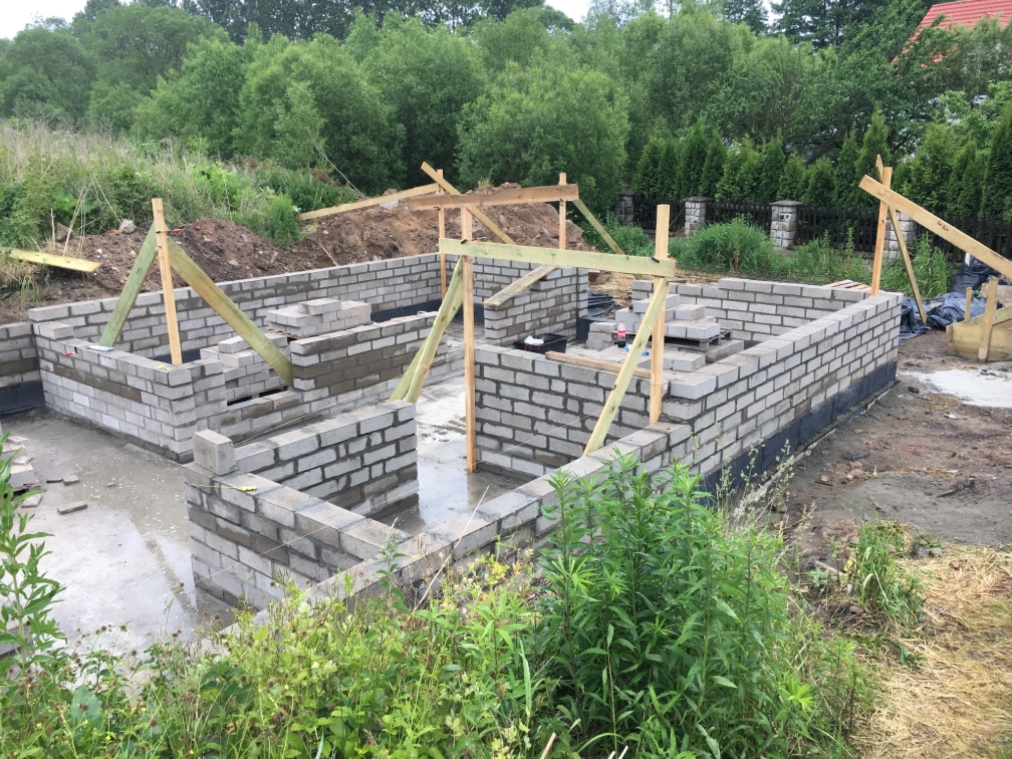 Fundamenty z bloczków betonowych, częściowo zalane wodą, z drewnianymi podporami i zielenią w tle.