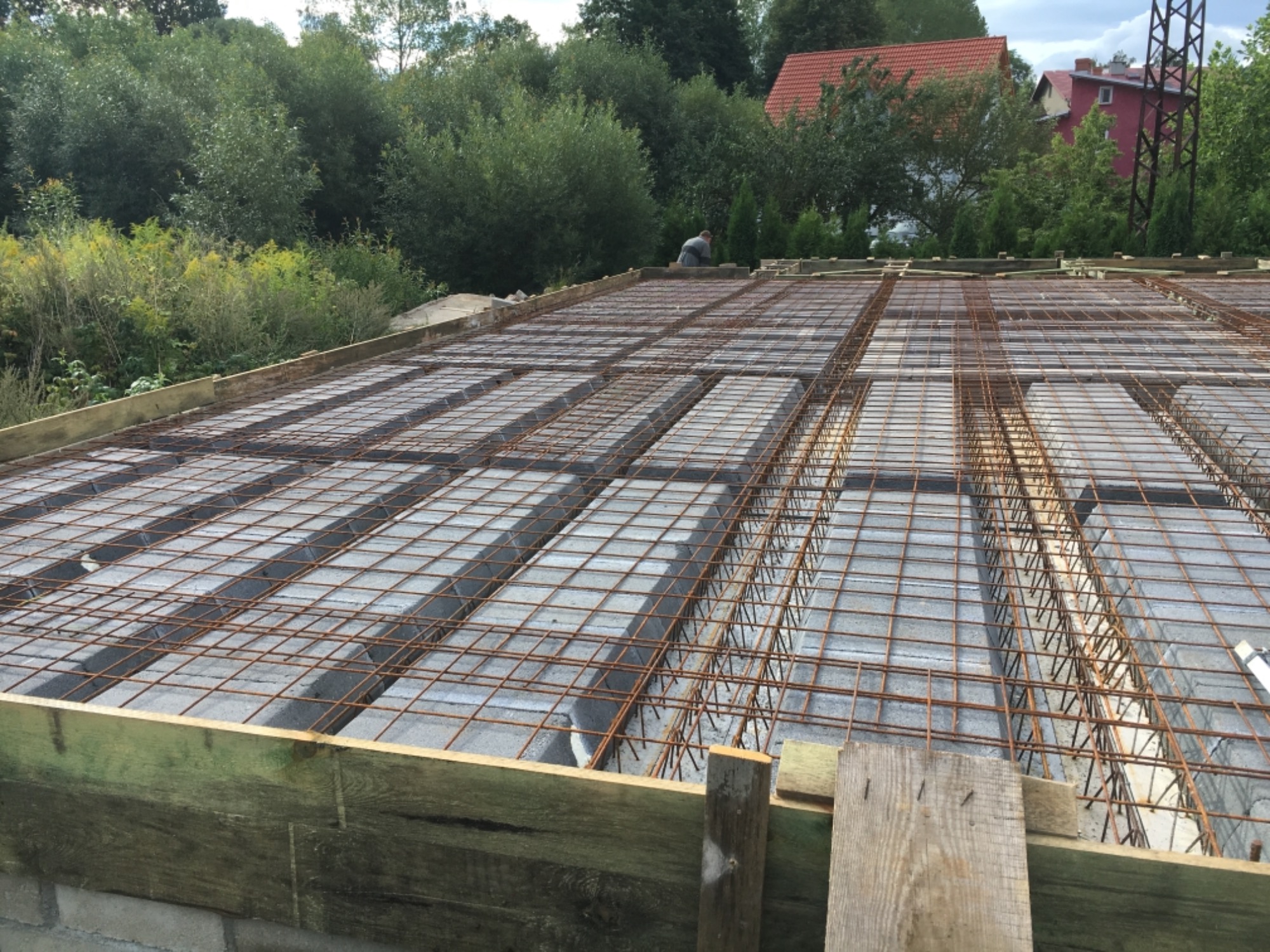 Zbrojenie stropu Teriva na budowie, widoczne bloczki betonowe, siatka zbrojeniowa i deski szalunkowe, w tle zieleń i dom z czerwoną dachówką.