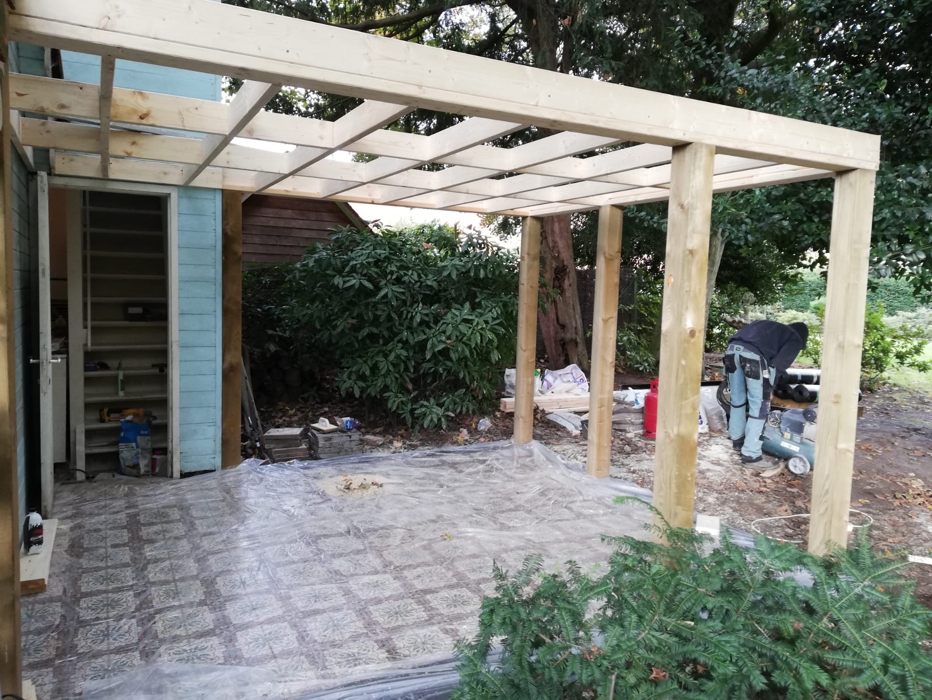 Konstrukcja drewnianej pergoli w trakcie budowy, widoczne słupy i kratownica, pracownik w roboczych spodniach i kompresor w tle, podłoże zabezpieczone folią.