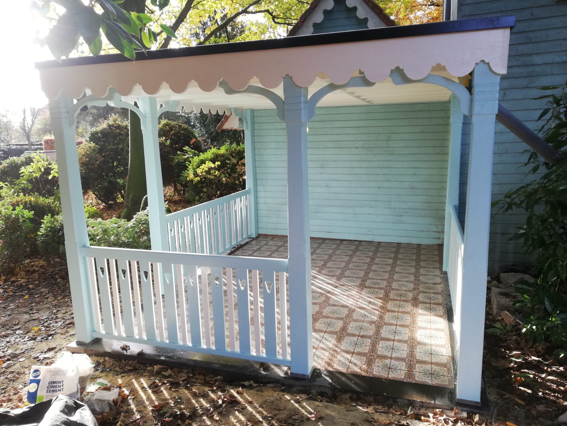 Świeżo wybudowana, ażurowa altana ogrodowa w odcieniach błękitu i bieli, z dekoracyjnym wykończeniem dachu i wzorzystą podłogą z płytek, obok worki z cementem.