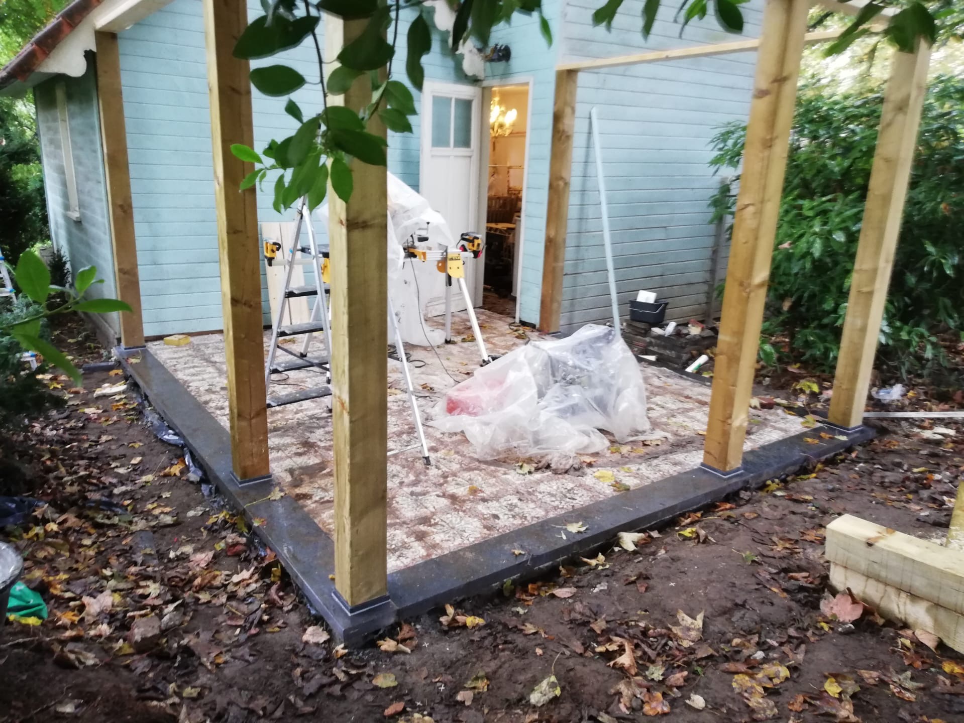 Budowa drewnianej konstrukcji altany z widocznym zadaszonym wejściem do budynku z jasnoniebieską elewacją, narzędzia oraz folia ochronna na podłożu z kostki.