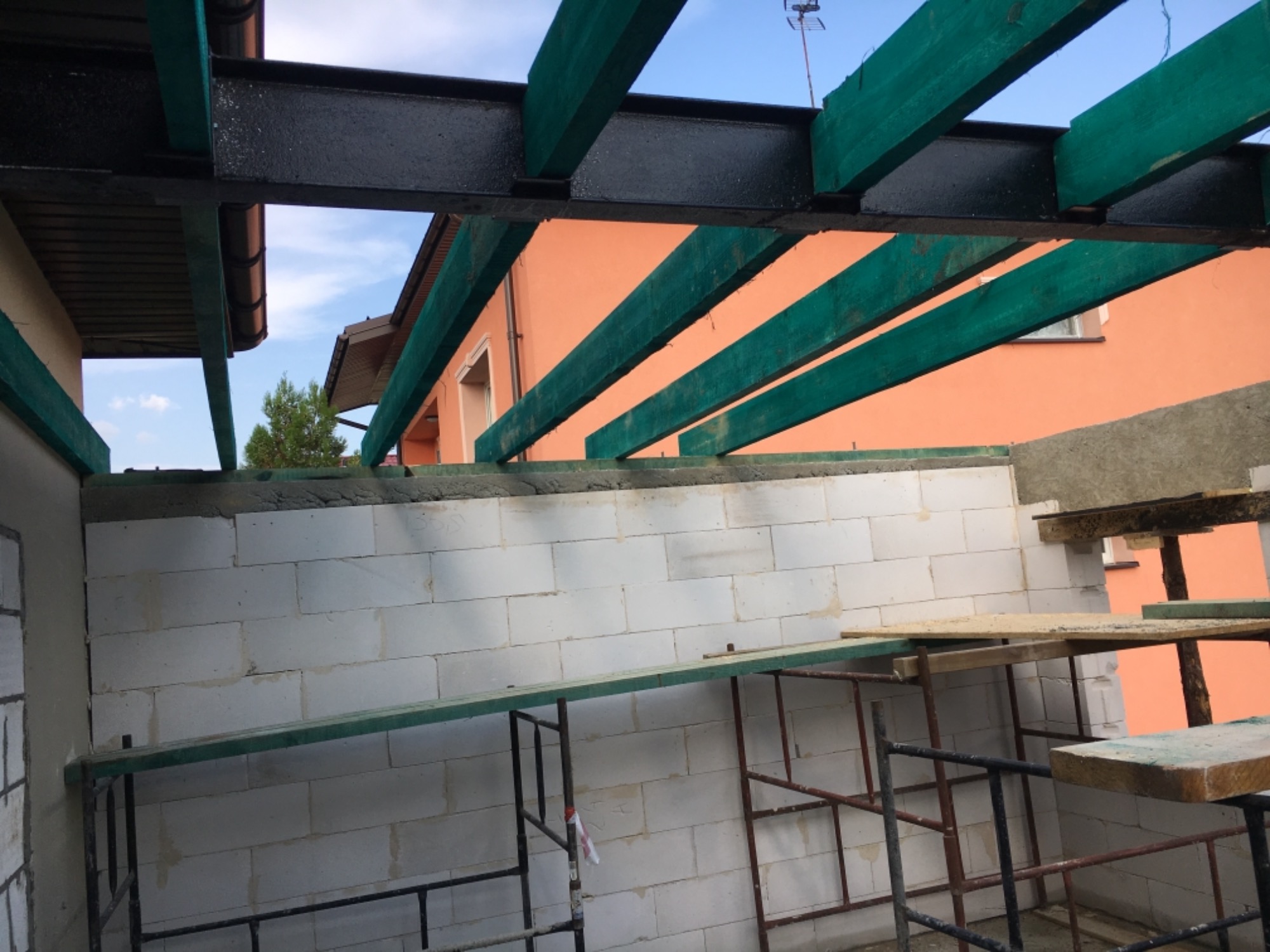 Wznoszenie ścian z betonu komórkowego na rusztowaniu, widoczne elementy konstrukcji dachu z belek drewnianych pomalowanych na zielono i stalowych belek na czarno.