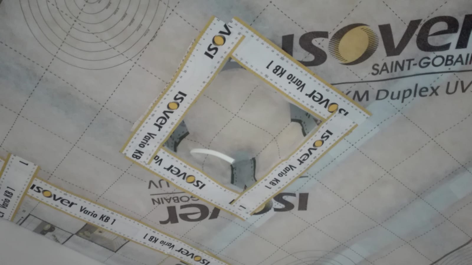 Fragment folii paroizolacyjnej Isover Vario KB 1 z wyciętym otworem, widoczne linie pomocnicze i logo producenta.