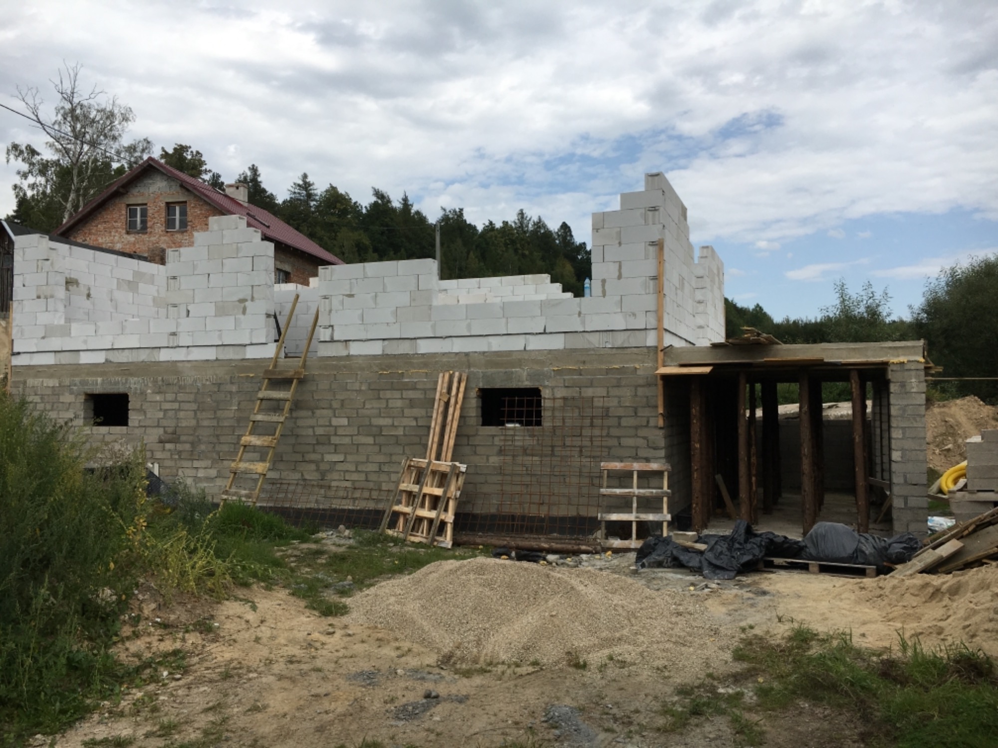 Budowa domu w stanie surowym otwartym: ściany z betonu komórkowego, widoczna drabina oparta o mur, sterta piasku i drewniane palety na placu budowy.