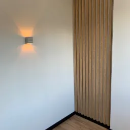 Drewniane lamele dekoracyjne na ścianie, oświetlenie punktowe, podłoga z paneli w odcieniach brązu, czarne listwy przypodłogowe.