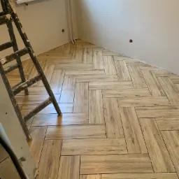 Ułożona podłoga z płytek imitujących drewno w jodełkę, widoczna drabina malarska z resztkami farby i fragment ściany z gniazdkiem elektrycznym.