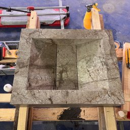 MAR-REM Usługi Remontowo Budowlane - Wykonana z kamienia umywalka na drewnianym stelażu, widoczna z góry. W tle narzędzia glazurnicze, w tym przecinarka do płytek i spryskiwacz. Umywalka o fakturze marmuru.