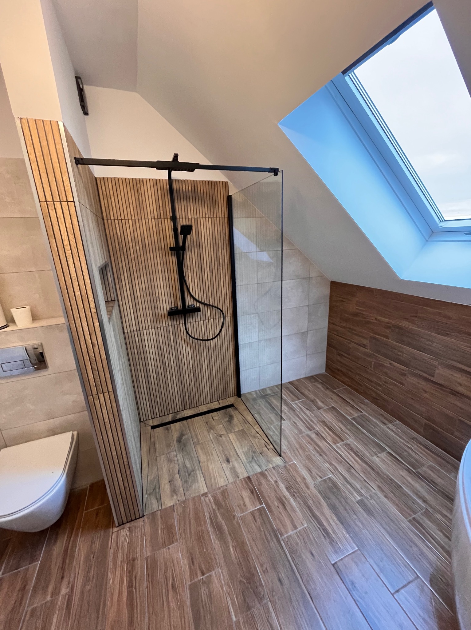 Nowoczesna kabina prysznicowa z drewnianymi panelami i czarną armaturą w łazience na poddaszu z oknem dachowym. Ułożone płytki imitujące drewno na podłodze i ścianach.