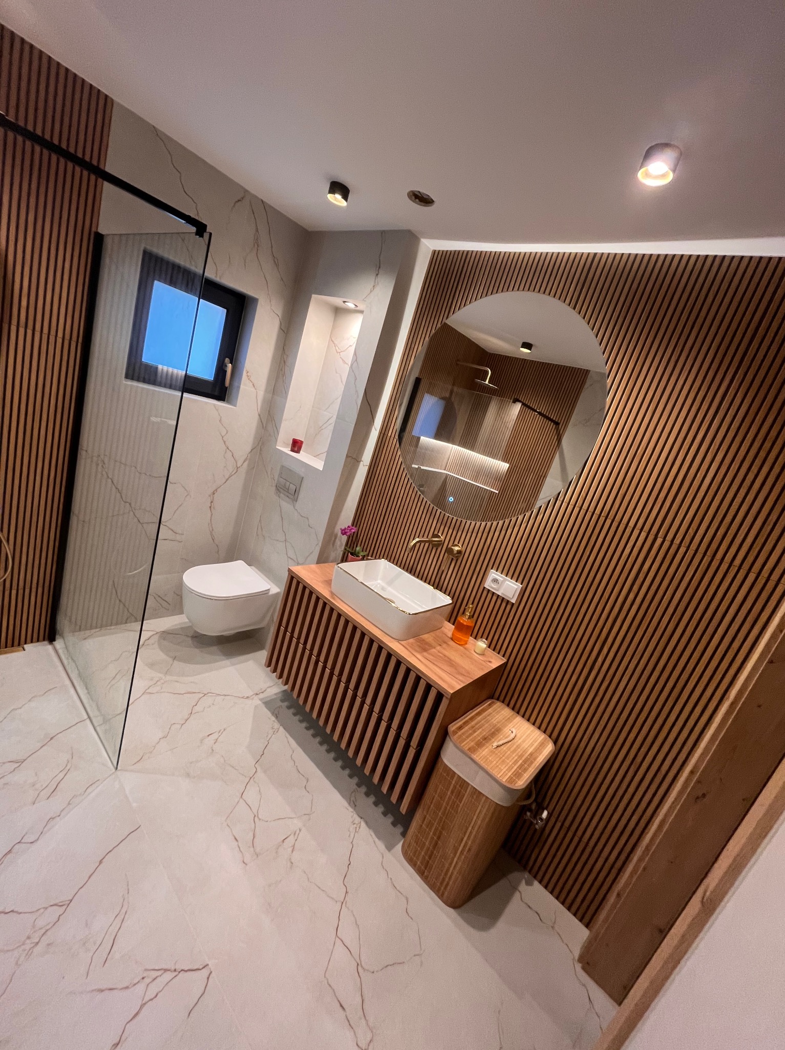 Nowoczesna łazienka z kabiną walk-in, umywalką nablatową na szafce z lamelami, okrągłym lustrem i marmurową płytką. Minimalistyczny design z drewnianymi akcentami.