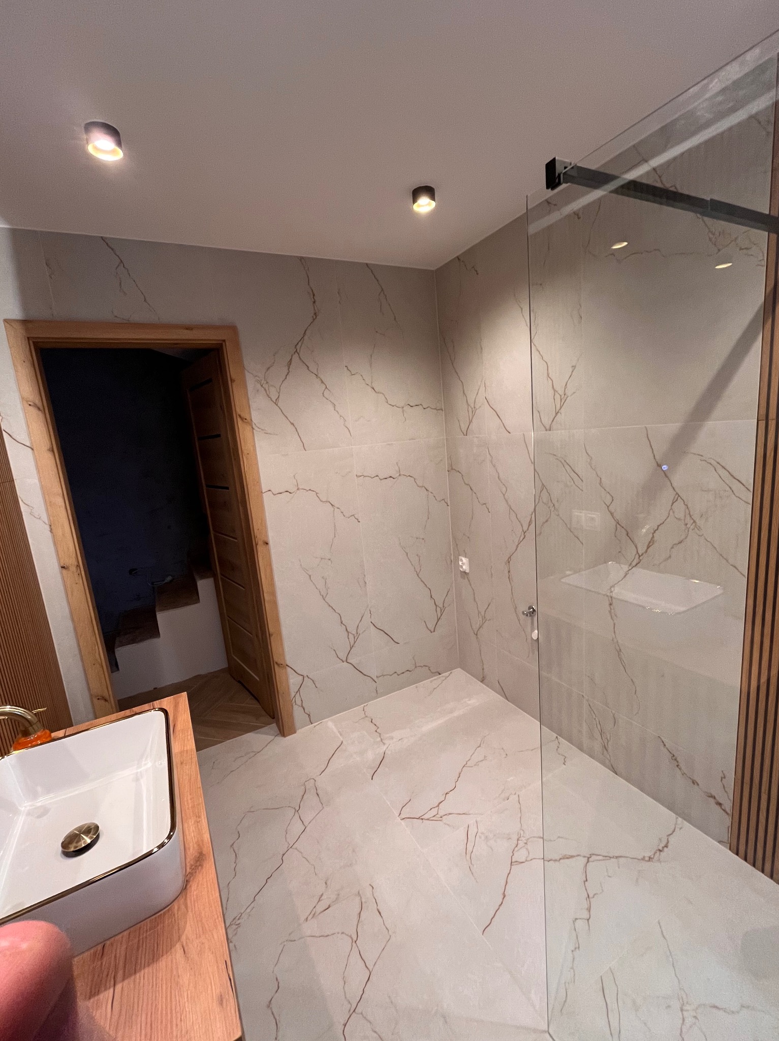 Nowoczesna łazienka z kabiną prysznicową typu walk-in, płytkami imitującymi marmur z brązowymi żyłami i drewnianymi akcentami. Widoczna umywalka nablatowa i oświetlenie punktowe.