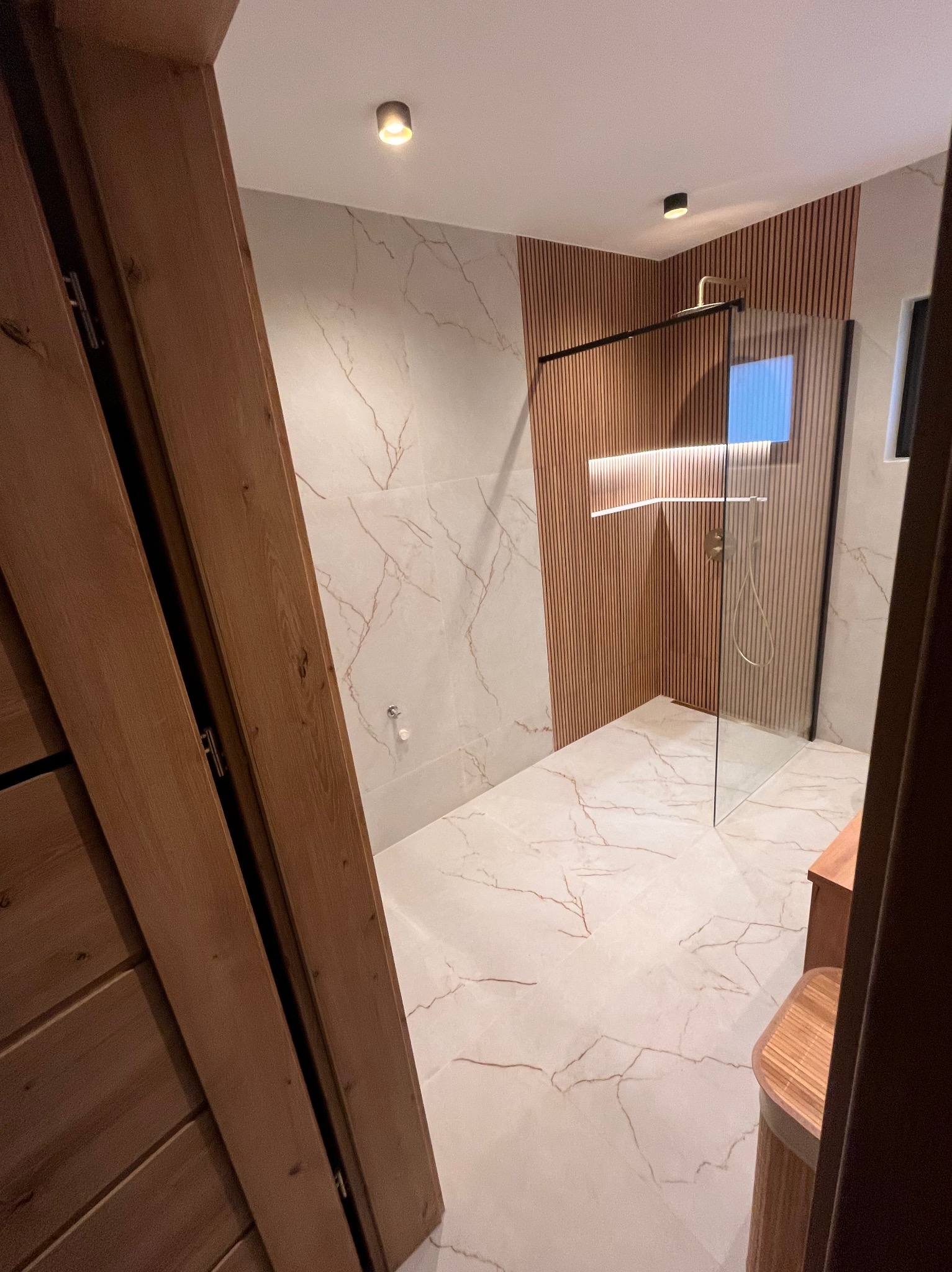 Nowoczesna łazienka z kabiną prysznicową typu walk-in, wykończona płytkami imitującymi marmur i drewnianymi panelami ściennymi. Widok zza drewnianych drzwi.