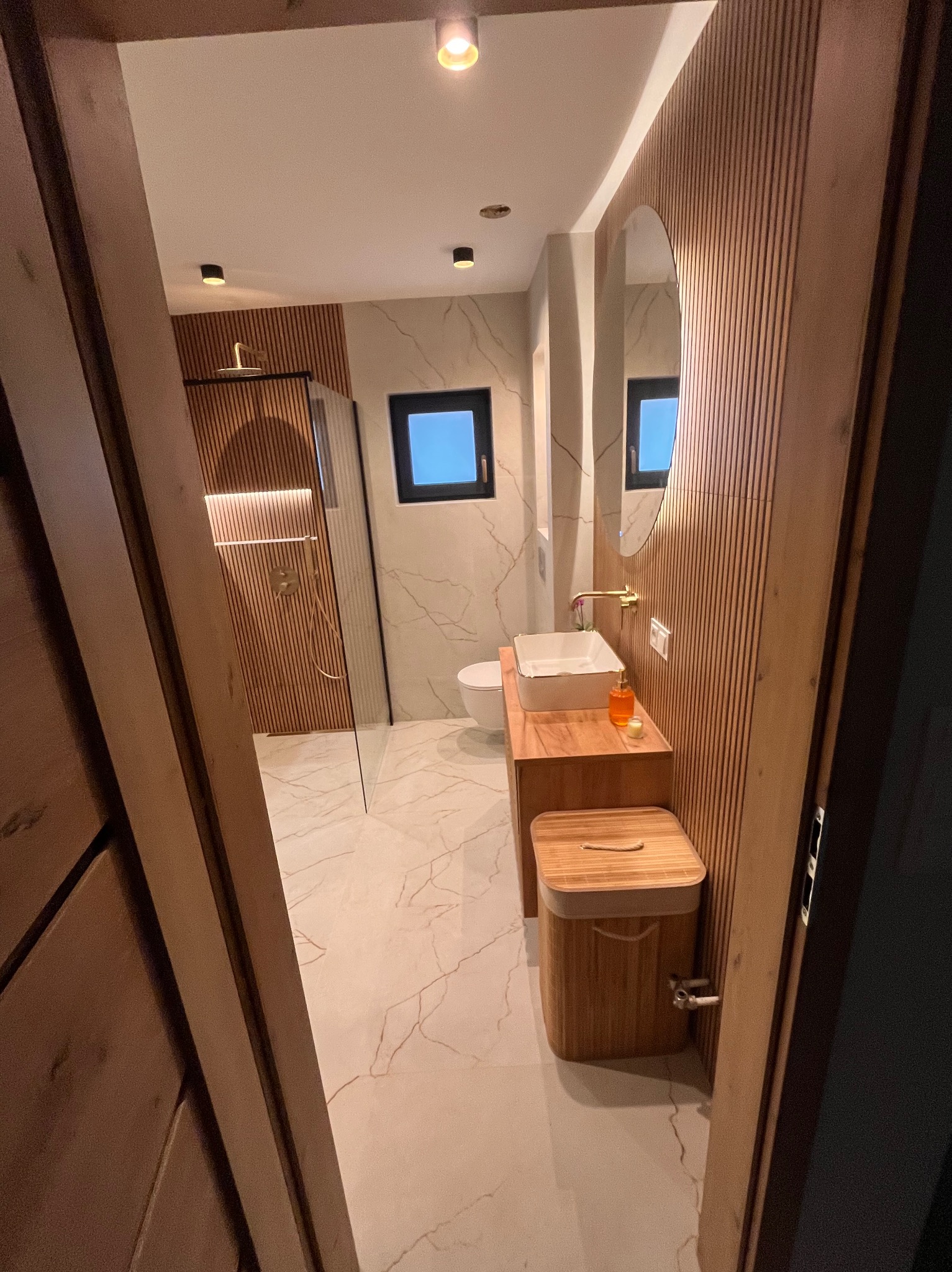 Nowoczesna łazienka z drewnianymi panelami ściennymi, marmurową podłogą i kabiną prysznicową typu walk-in. Umywalka nablatowa i okrągłe lustro dodają elegancji.