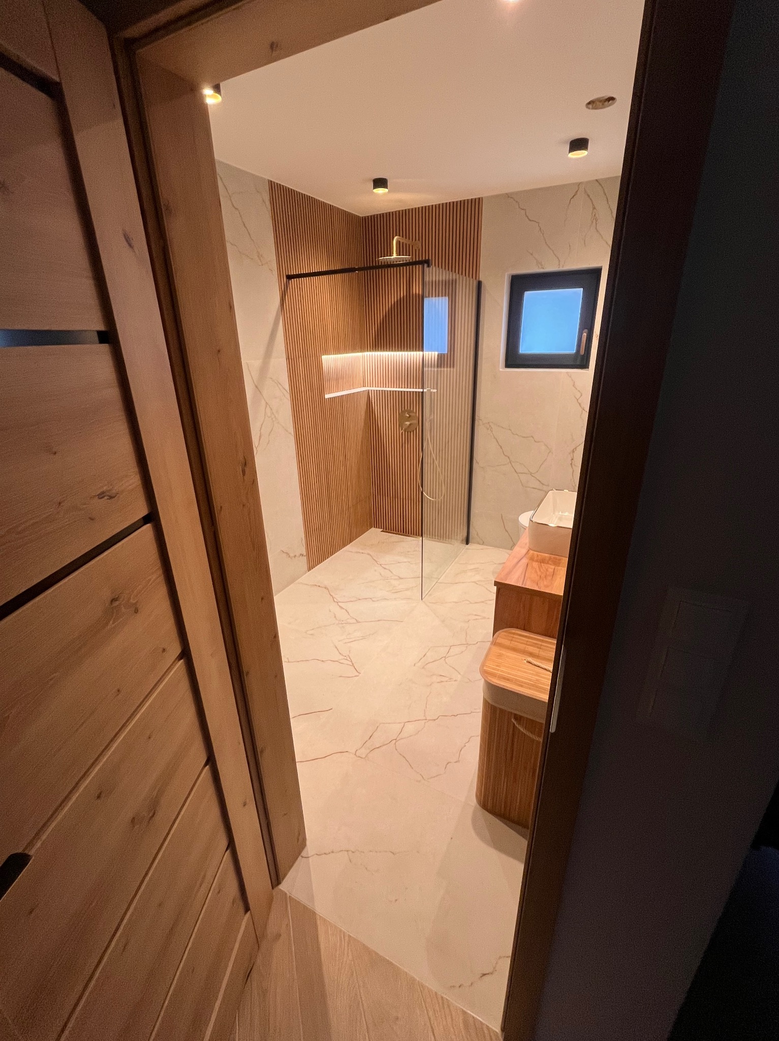 Nowoczesna łazienka z drewnianymi akcentami: kabina prysznicowa ze złotą armaturą, marmurowe płytki i drewniane panele ścienne, widok zza otwartych drzwi.