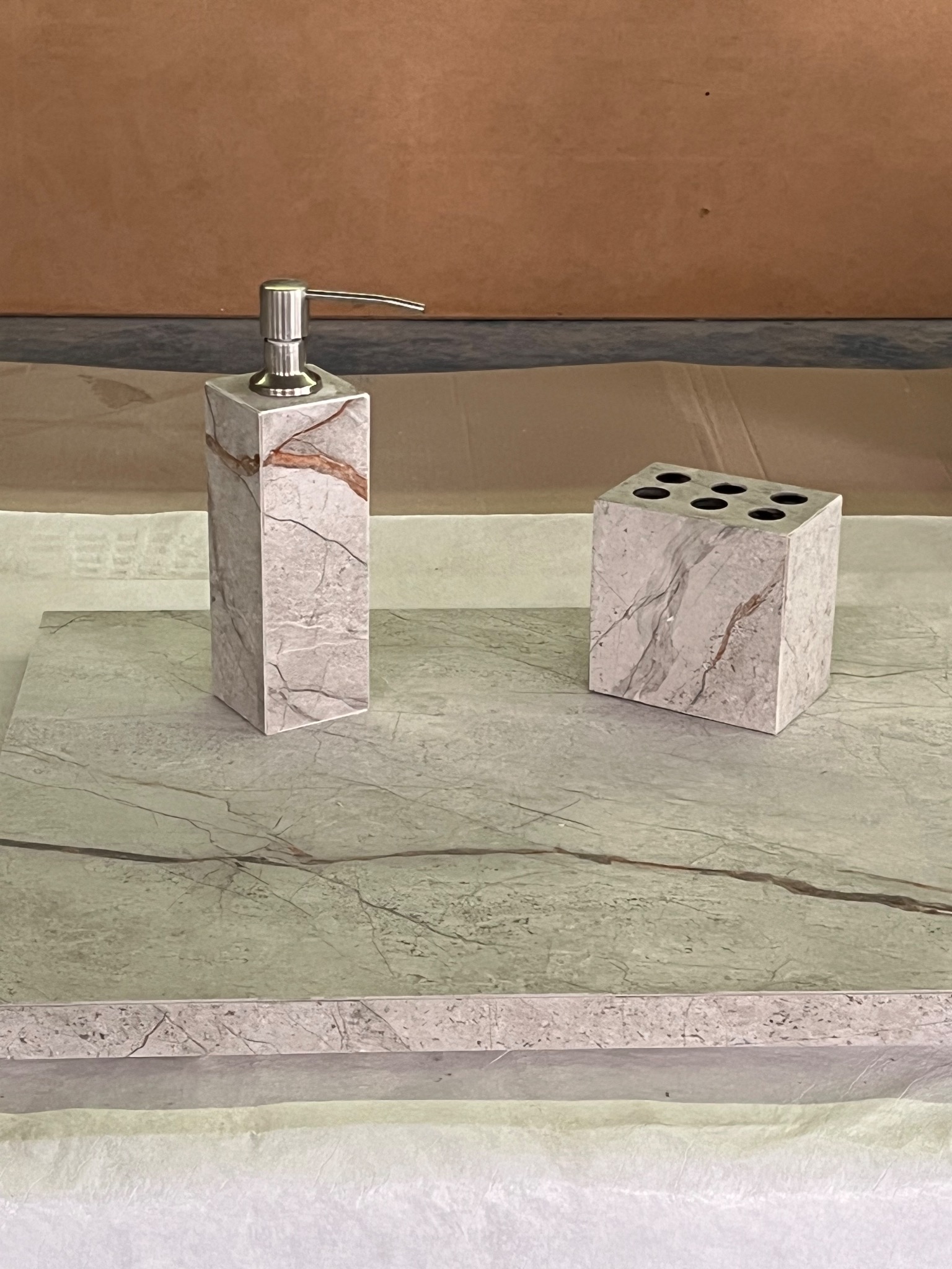 Marmurowy dozownik mydła, kubek na szczoteczki i podkładka, zestaw łazienkowy w neutralnych barwach, z widoczną fakturą kamienia i metalowym aplikatorem.