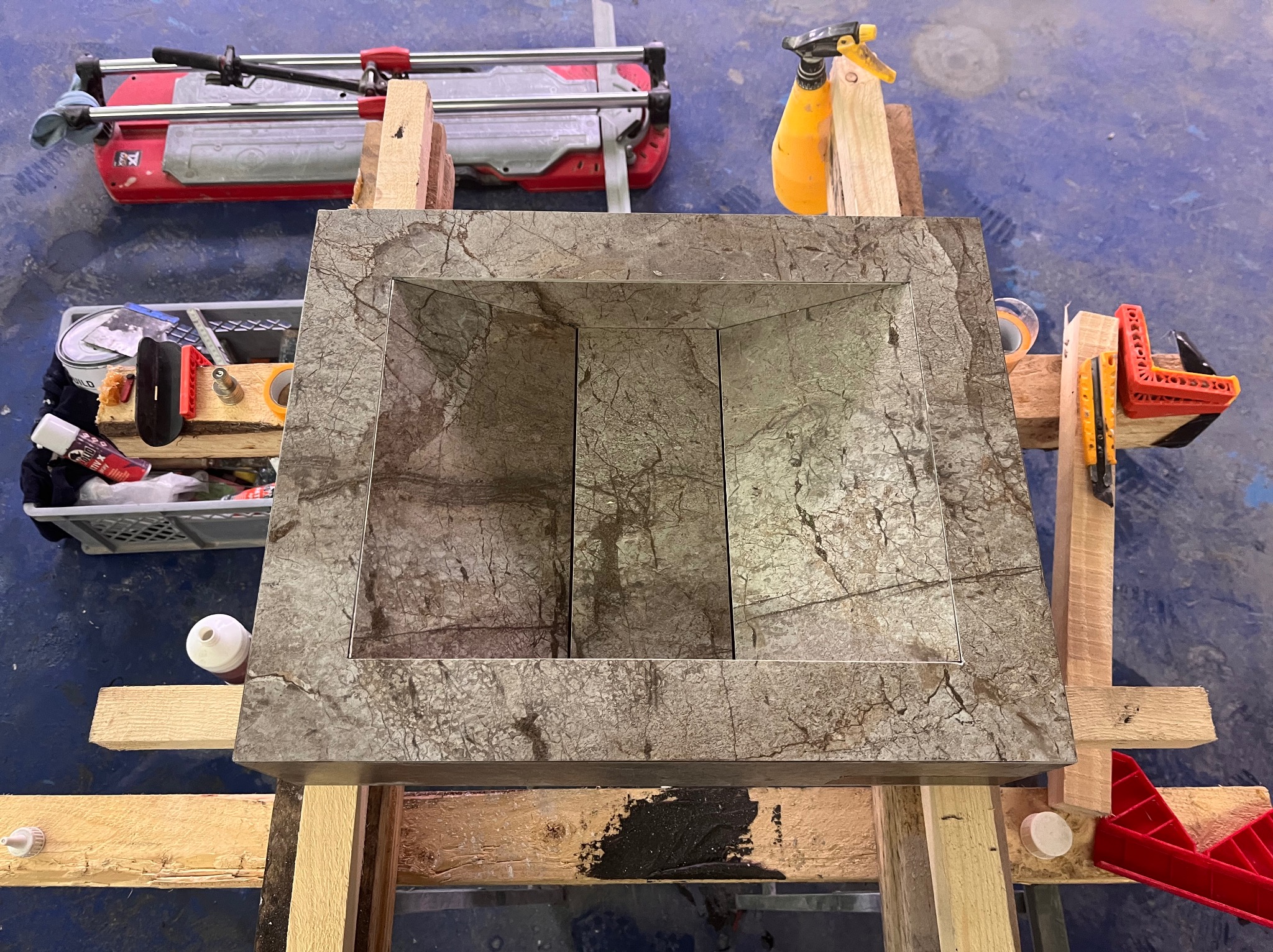 Wykonana z kamienia umywalka na drewnianym stelażu, widoczna z góry. W tle narzędzia glazurnicze, w tym przecinarka do płytek i spryskiwacz. Umywalka o fakturze marmuru.