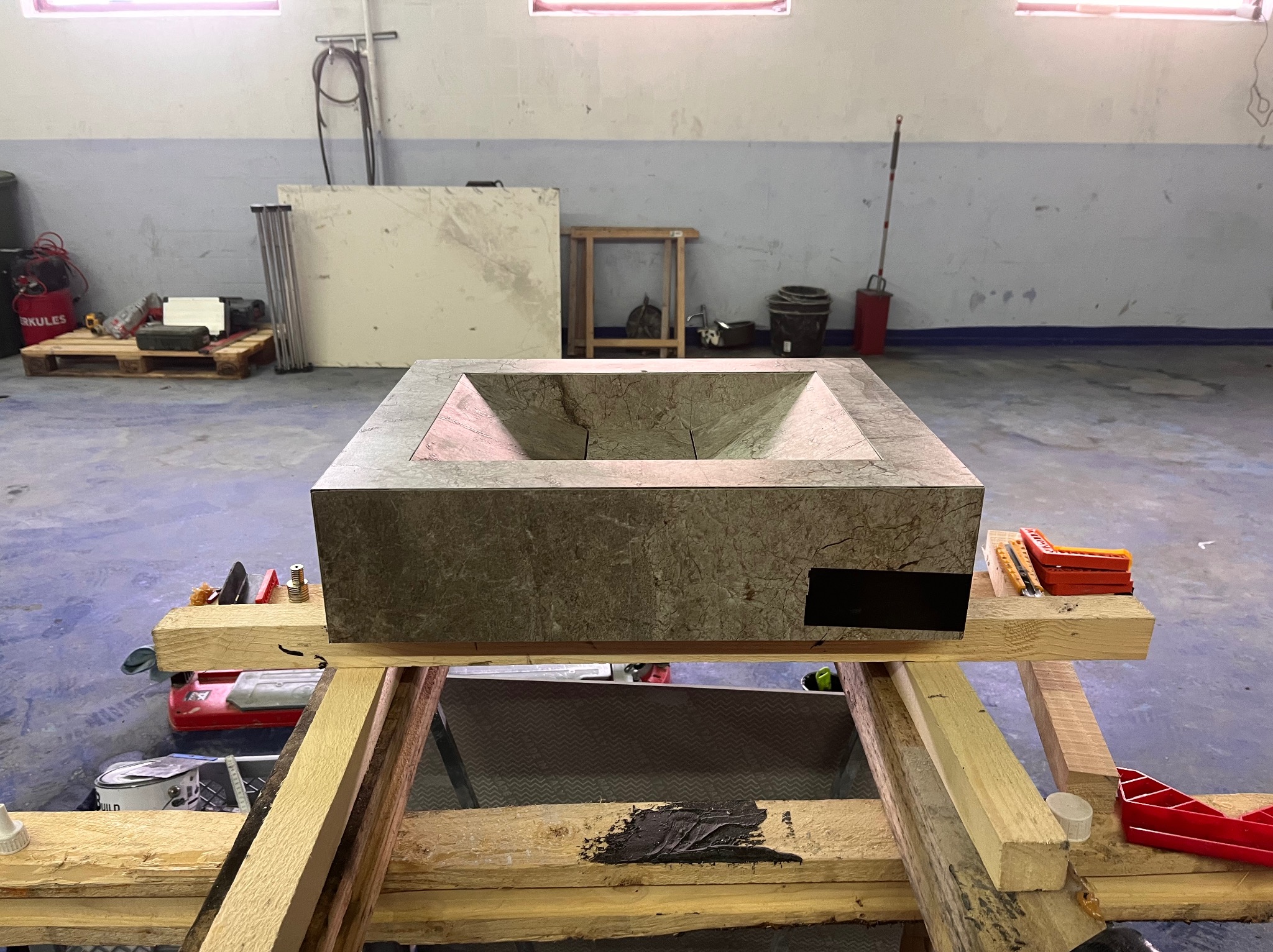 Kamienna umywalka w kształcie prostopadłościanu, umieszczona na drewnianym stelażu w warsztacie. Widoczne narzędzia i materiały budowlane w tle. Surowy, industrialny charakter.