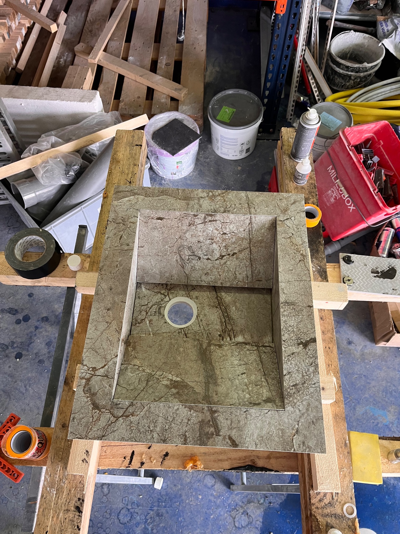 Marmurowa umywalka w trakcie tworzenia, wsparta na drewnianej konstrukcji. Widoczne narzędzia i materiały budowlane w tle. Ujęcie z góry, podkreślające detale kamienia.