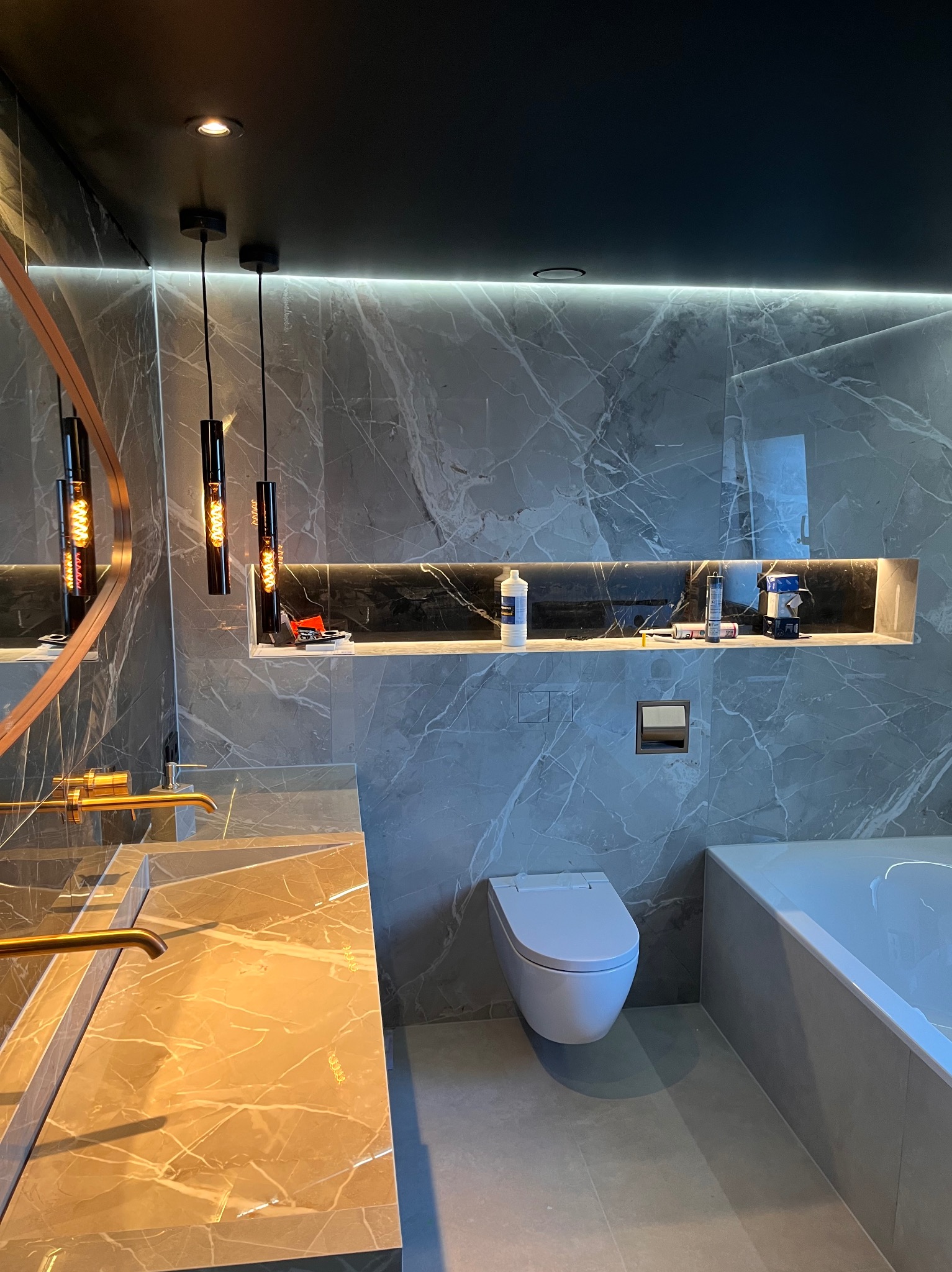 Elegancka łazienka z marmurowymi płytkami, złotą armaturą i podświetlaną wnęką. Nowoczesny design z wiszącą toaletą i minimalistycznym oświetleniem.