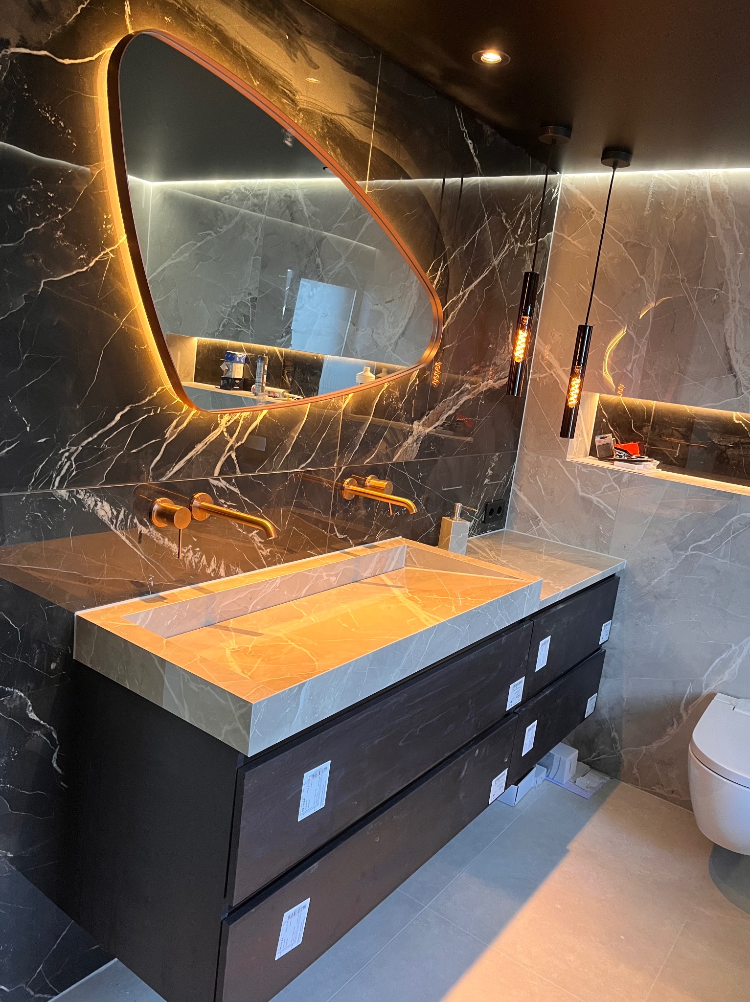 Luksusowa łazienka z marmurowymi ścianami, podświetlanym lustrem o nieregularnym kształcie i nowoczesną umywalką z szafką w ciemnym odcieniu drewna.