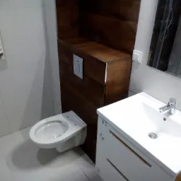 Nowoczesna łazienka po remoncie: podwieszana toaleta z zabudową w odcieniach drewna, biała umywalka z szafką i bateria chromowana, białe płytki na ścianach i podłodze, widoczny grzejnik drabinkowy.