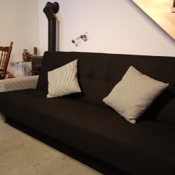 Pracownia Tapicerska Karol Krupa - Czarna sofa z dwoma poduszkami w pepitkę, drewniane schody w tle, widoczny piec.