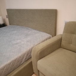 Pracownia Tapicerska Karol Krupa - Łóżko tapicerowane z pikowanym wezgłowiem w odcieniach beżu i fotel tapicerowany w tym samym kolorze, obok łóżka.