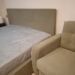 Łóżko tapicerowane z pikowanym wezgłowiem w odcieniach beżu i fotel tapicerowany w tym samym kolorze, obok łóżka.