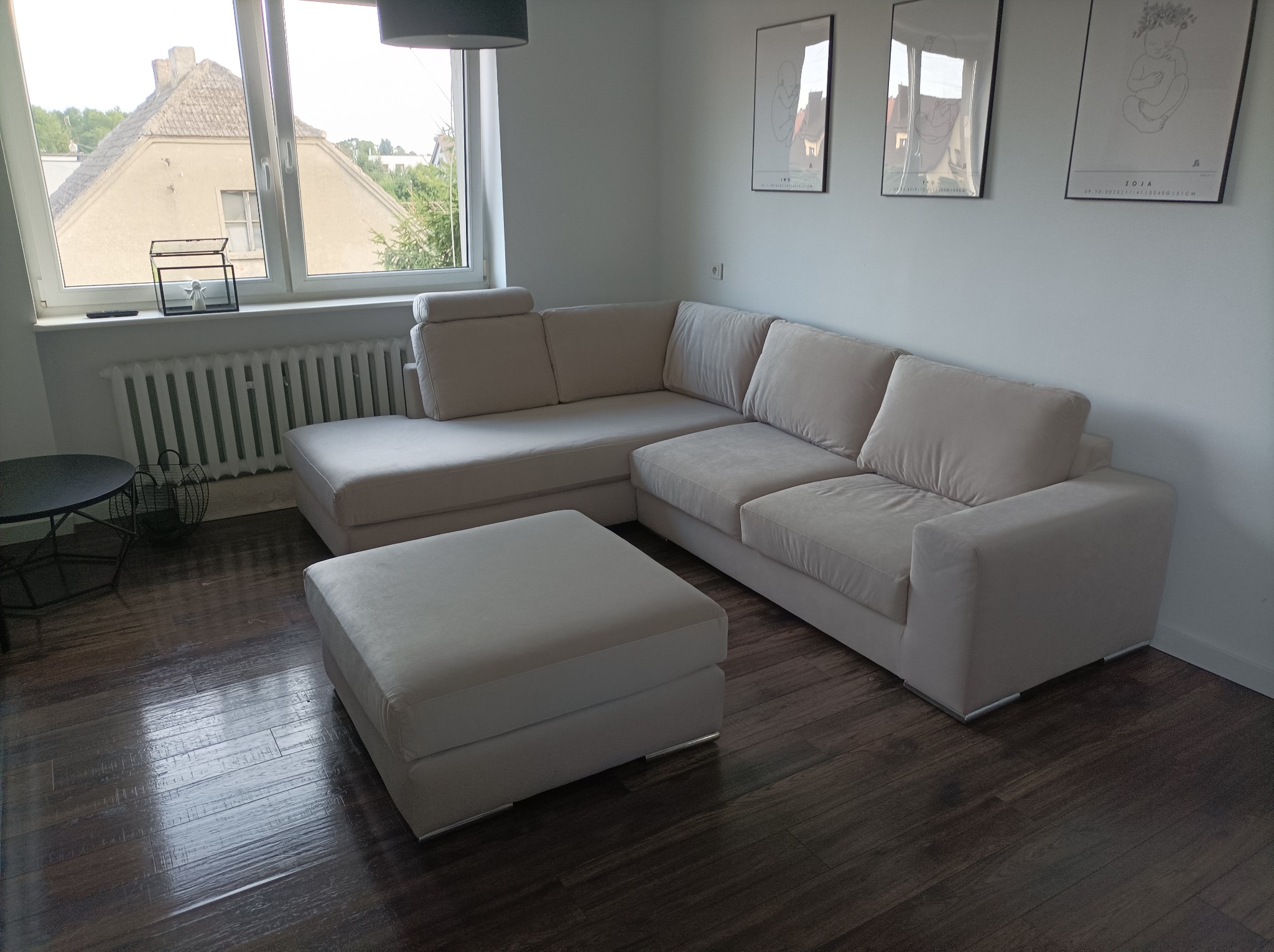 Jasny narożnik z poduszkami i pufą na ciemnej, drewnianej podłodze w salonie z trzema obrazami na ścianie.