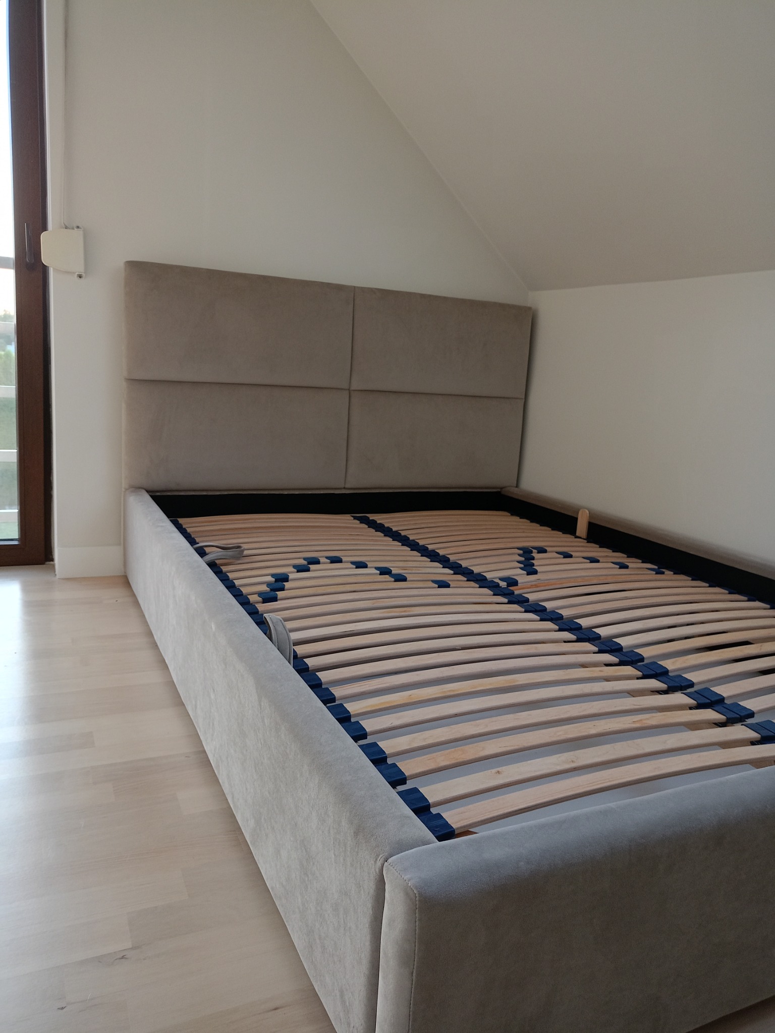 Beżowe tapicerowane łóżko z drewnianym stelażem listwowym, widok z niskiego kąta, jasne wnętrze.