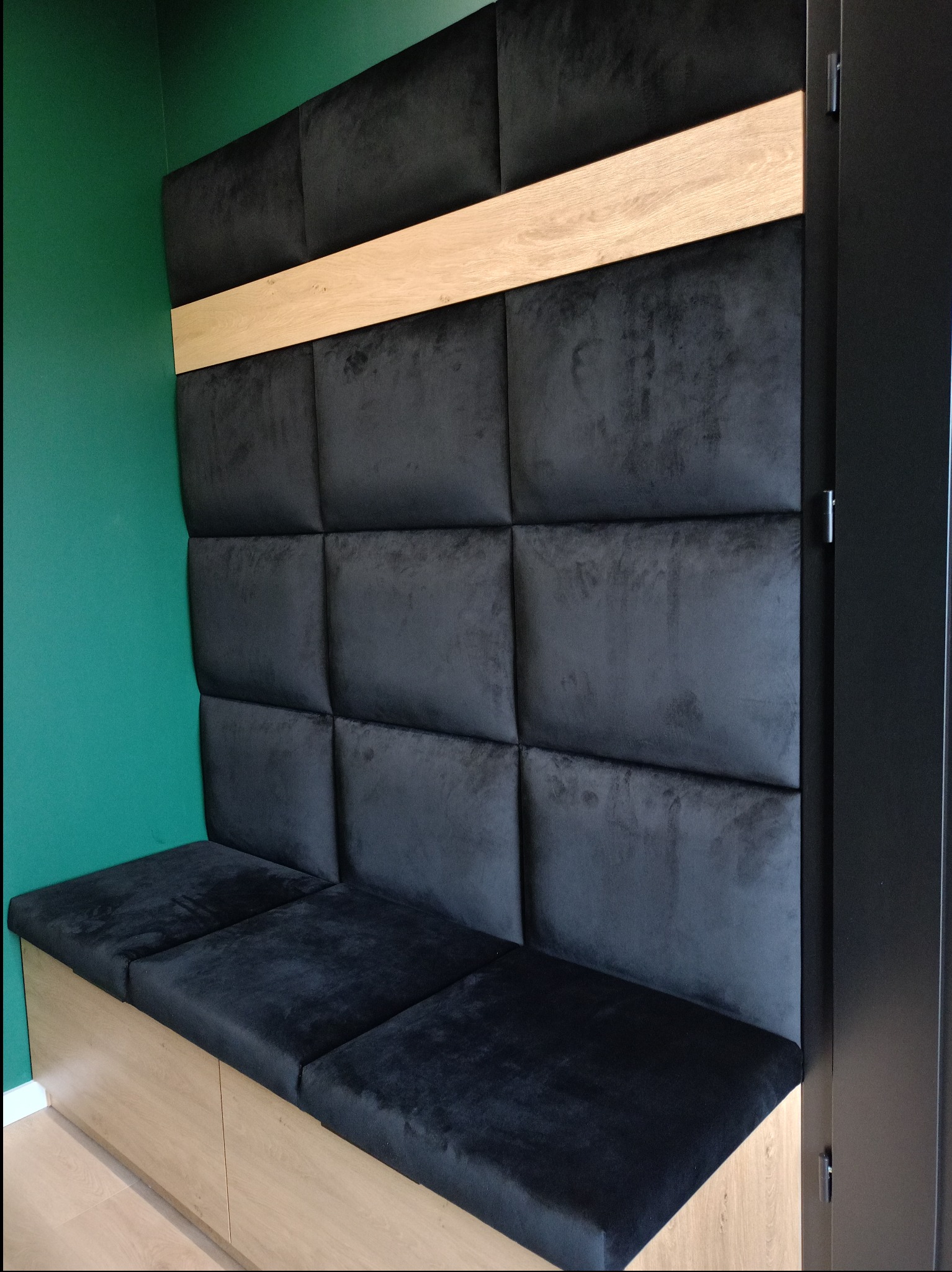 Wnęka z siedziskiem i tapicerowanym panelem ściennym w kolorze czarnym, z drewnianą listwą dekoracyjną i szafkami pod siedziskiem, na tle zielonej ściany.