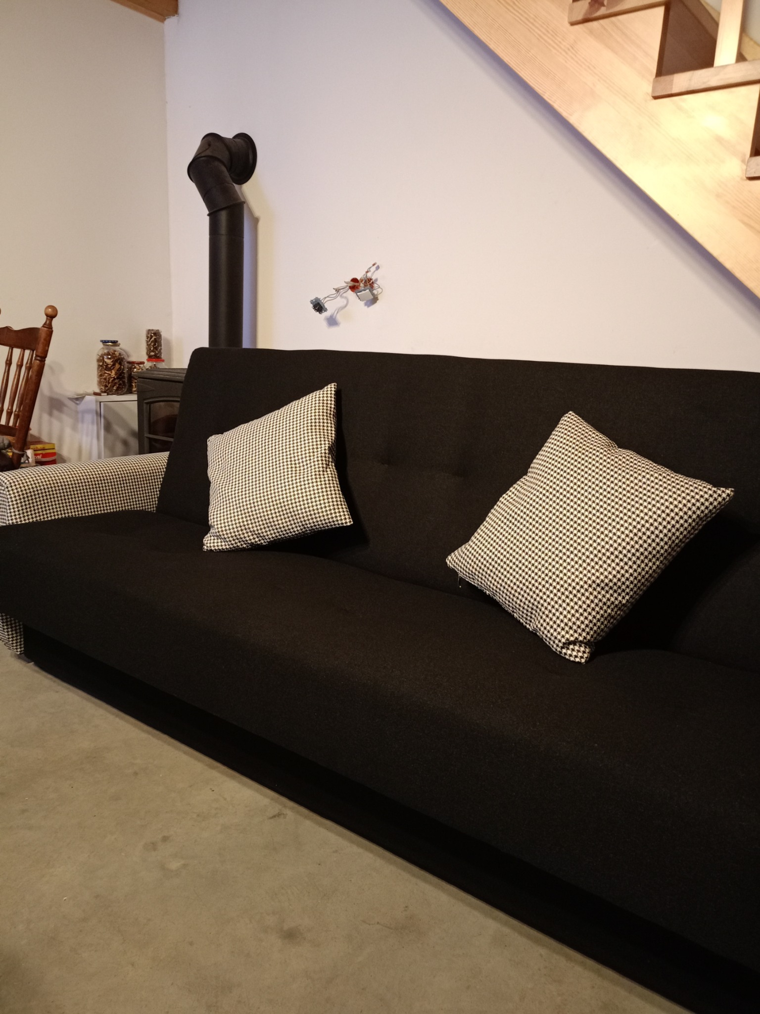 Czarna sofa z dwoma poduszkami w pepitkę, drewniane schody w tle, widoczny piec.