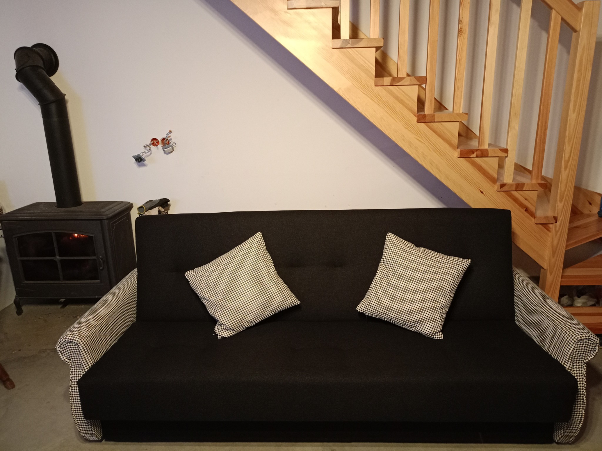 Czarna rozkładana sofa z poduszkami w pepitkę, ustawiona w pokoju z kominkiem i drewnianymi schodami.