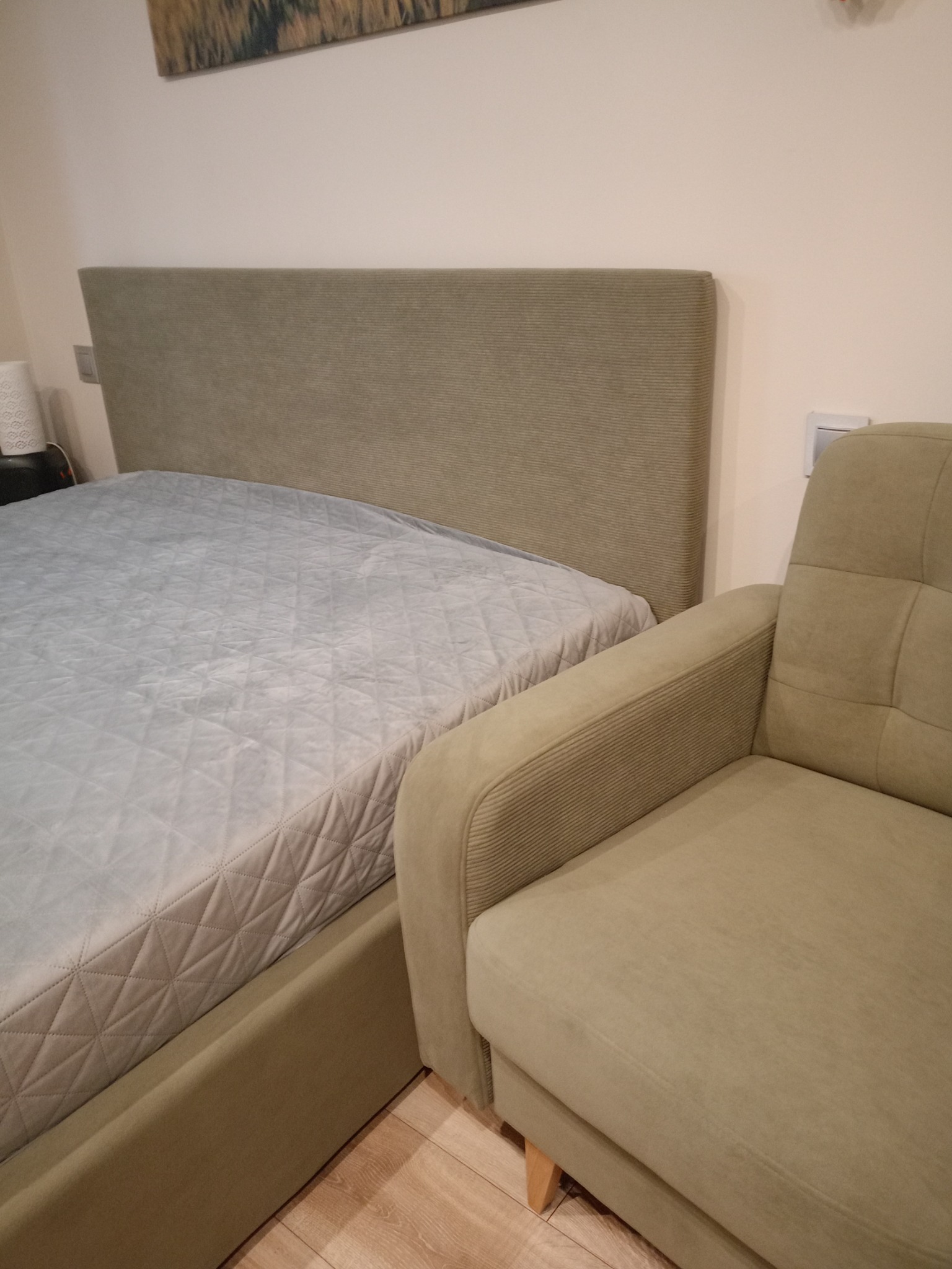 Łóżko tapicerowane z pikowanym wezgłowiem w odcieniach beżu i fotel tapicerowany w tym samym kolorze, obok łóżka.