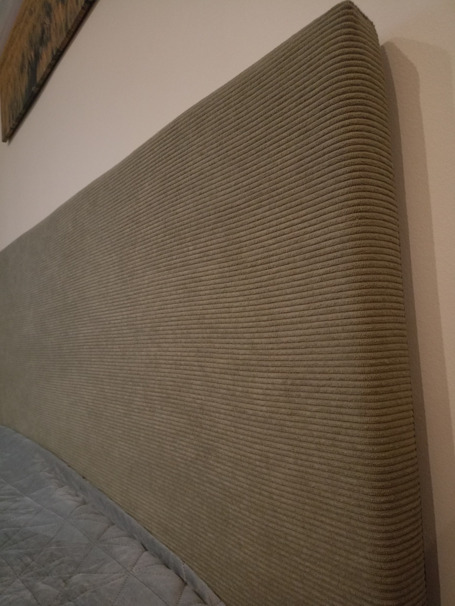 Beżowy, tapicerowany zagłówek łóżka z tkaniny w prążki, widoczny fragment pikowanej narzuty na łóżko i fragment obrazu na ścianie.