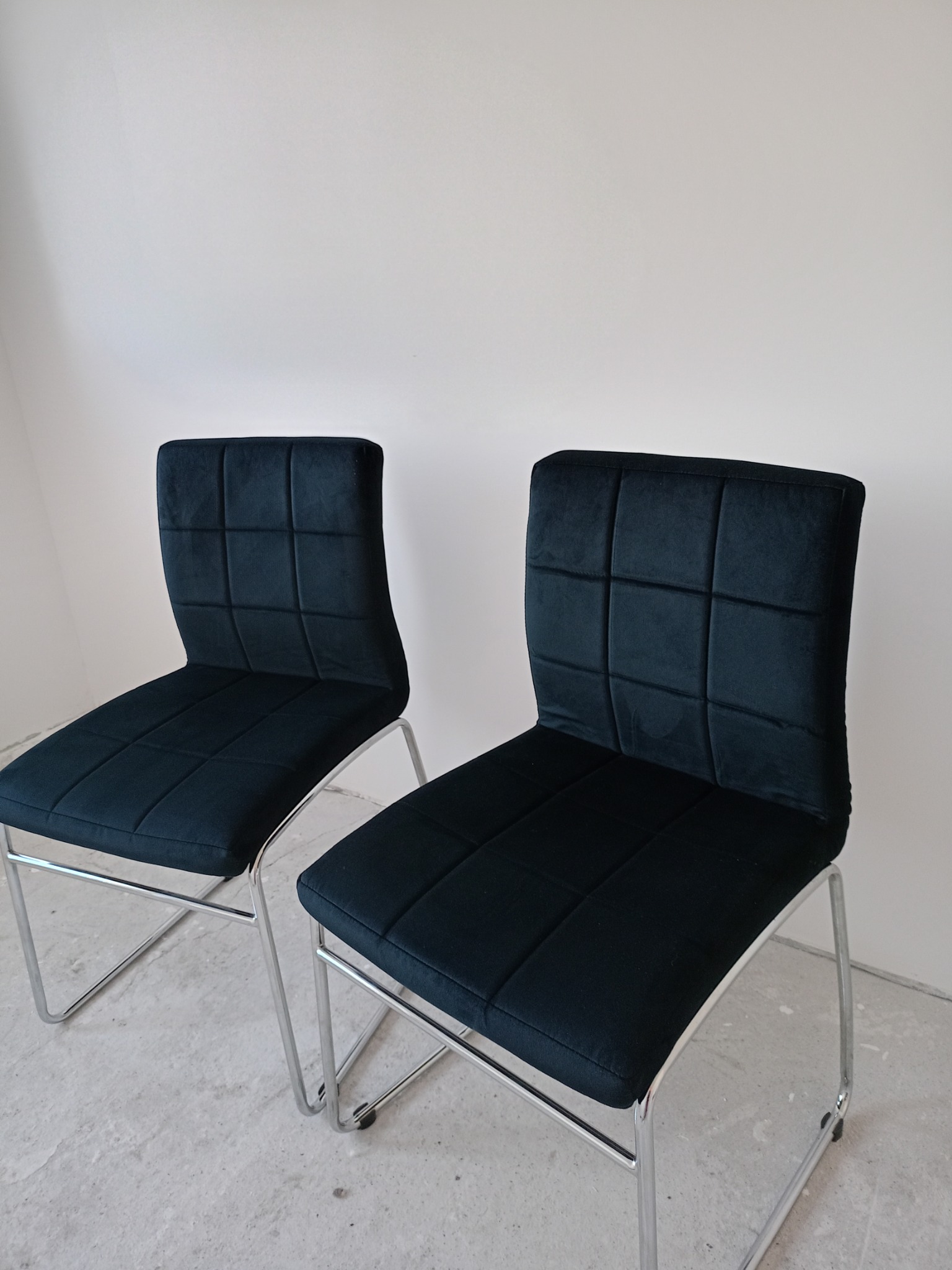 Dwa czarne krzesła tapicerowane z kwadratowymi przeszyciami na siedzisku i oparciu, z chromowanymi nogami, ustawione na szarej podłodze przy białej ścianie.