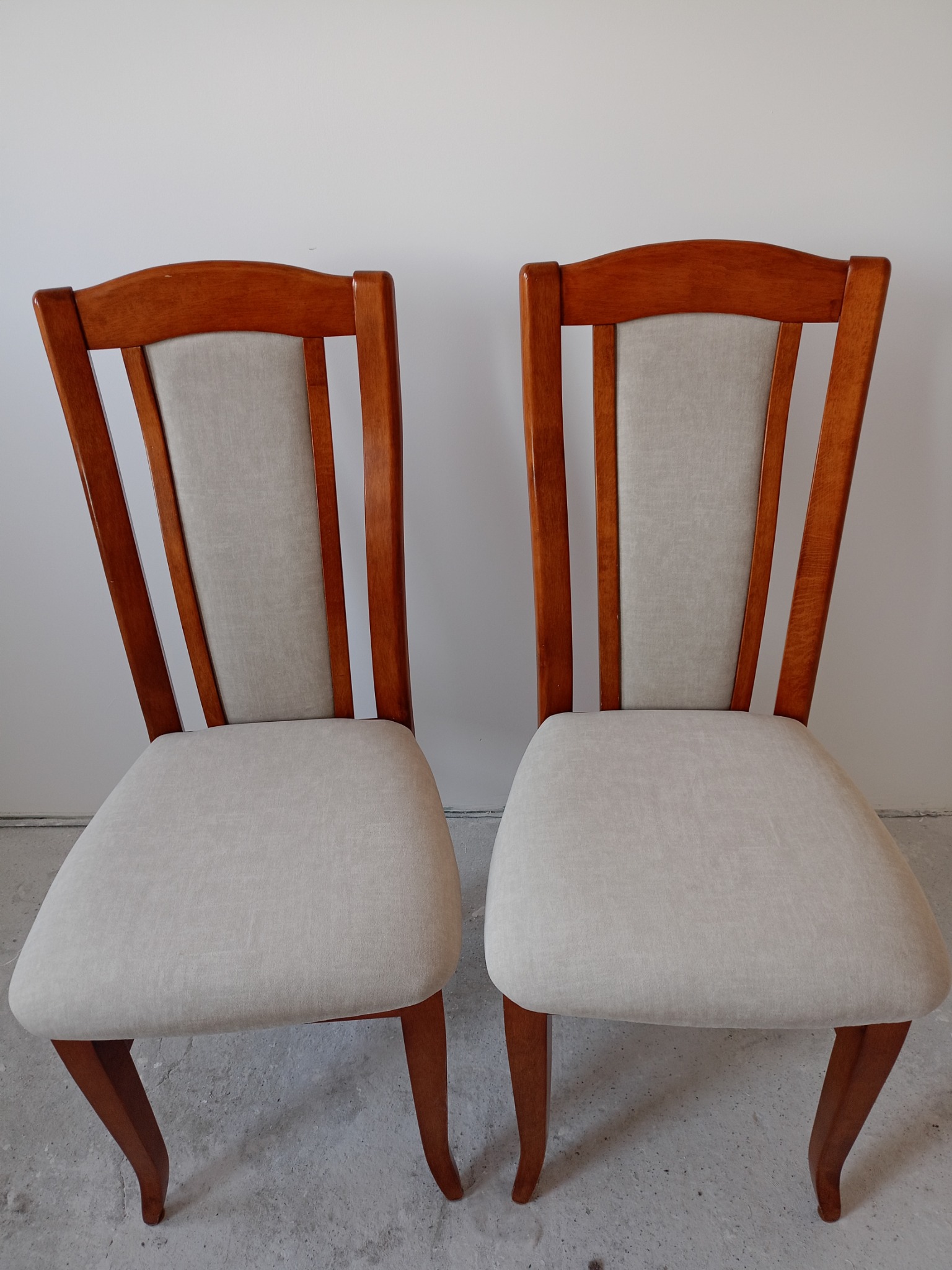 Dwa krzesła z drewnianą ramą w odcieniach brązu i jasną tapicerką, ustawione na szarym tle w minimalistycznym wnętrzu.