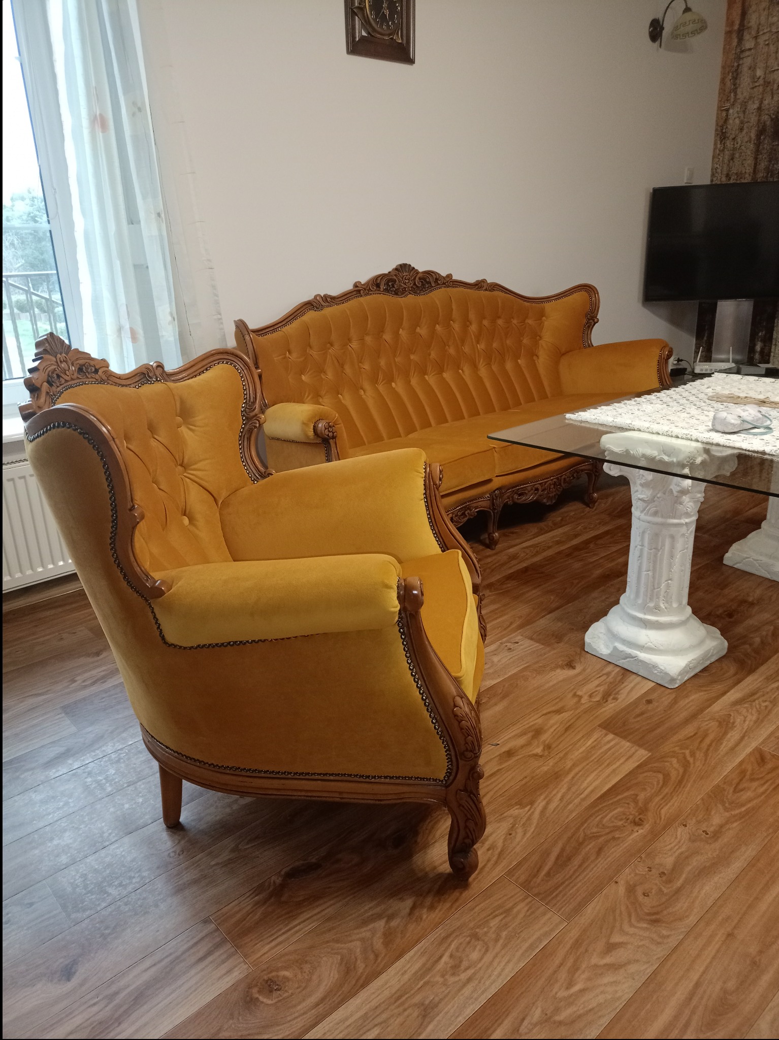 Żółty, tapicerowany fotel i sofa w stylu retro z drewnianymi zdobieniami, na drewnianej podłodze, obok stolika z blatem szklanym i kolumnowymi nogami.