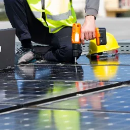 Instalator w kamizelce odblaskowej przykręca śrubokrętem panele słoneczne; obok leży kask i otwarty laptop.