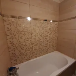 Beżowa łazienka z wanną, wykończona płytkami z motywem roślinnym i metalową listwą dekoracyjną, widok na baterię wannową z niebieskim elementem.