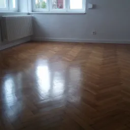 Wycyklinowany, lakierowany parkiet ułożony we wz&oacute;r jodełki klasycznej w pustym, jasnym pokoju z grzejnikiem pod oknem.