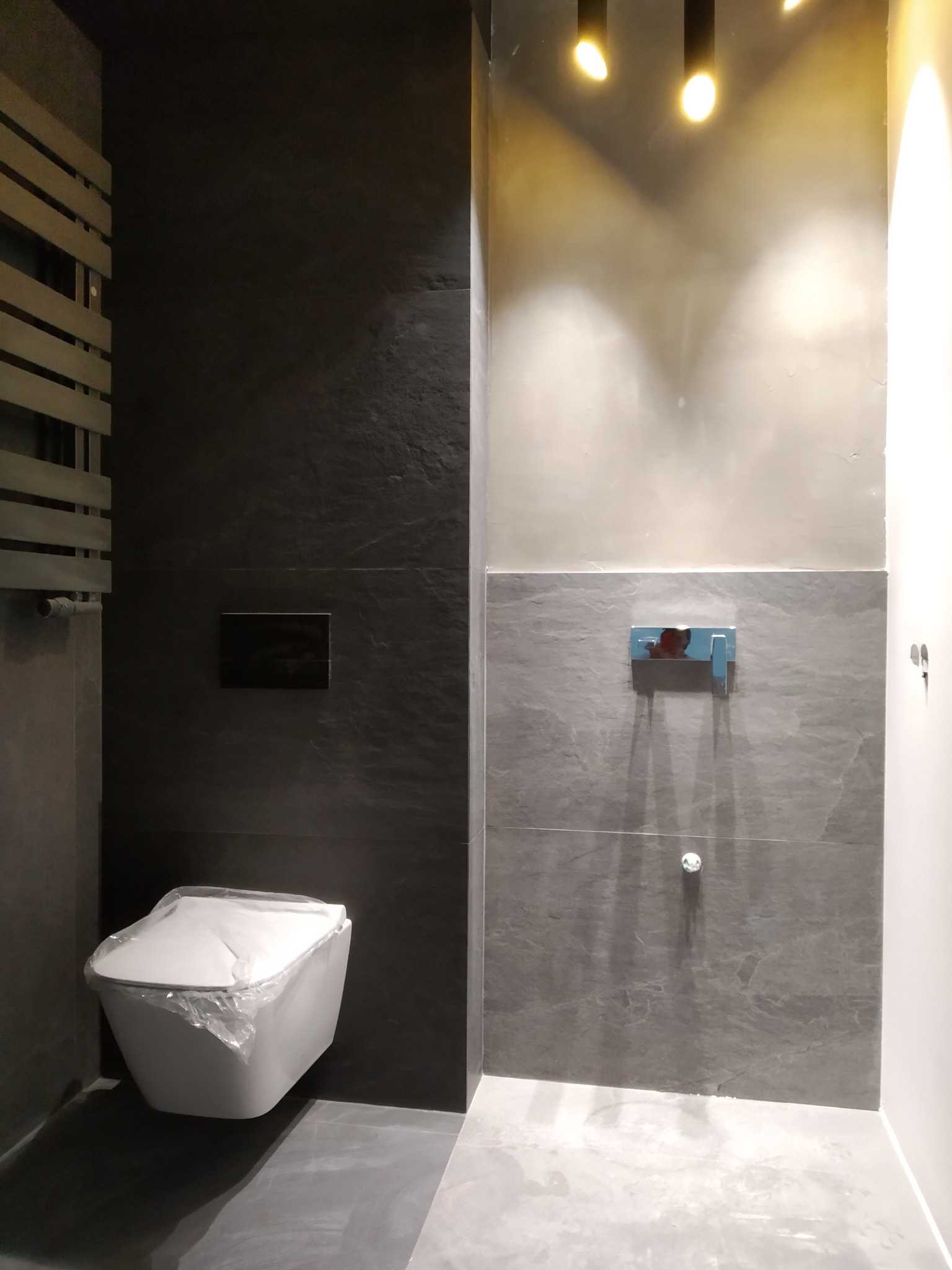 Nowoczesna łazienka w odcieniach szarości z wiszącą toaletą zabezpieczoną folią, widoczny grzejnik drabinkowy, bateria umywalkowa i oświetlenie punktowe.