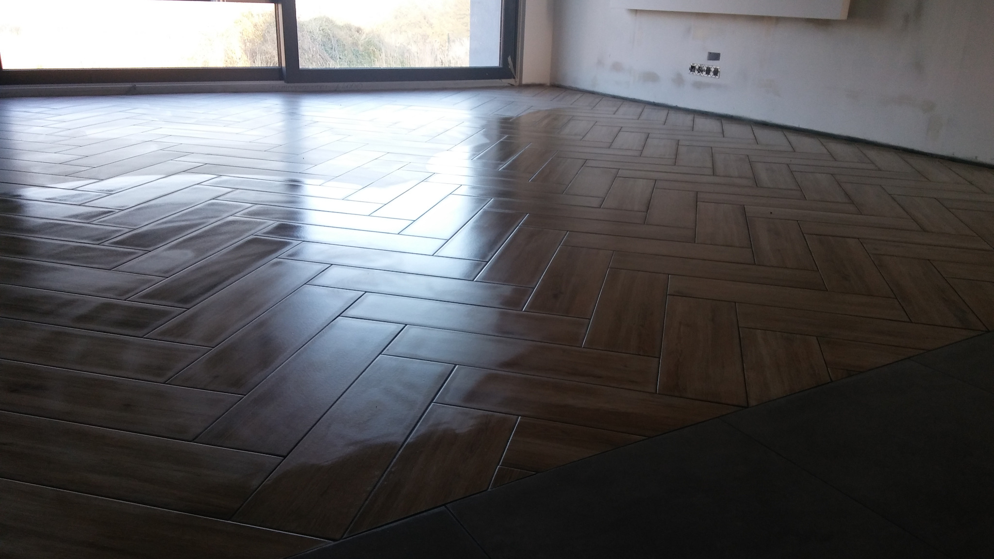 Ułożona w jodełkę, błyszcząca, brązowa podłoga z płytek imitujących drewno, widok na fragment ściany z gniazdkami elektrycznymi i okno w tle.