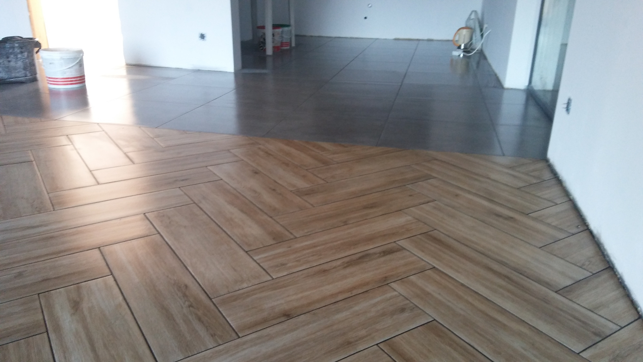 Ułożone płytki podłogowe w jodełkę, przechodzące w szare, gładkie płytki w tle, widoczne białe ściany i wiaderka z farbą.