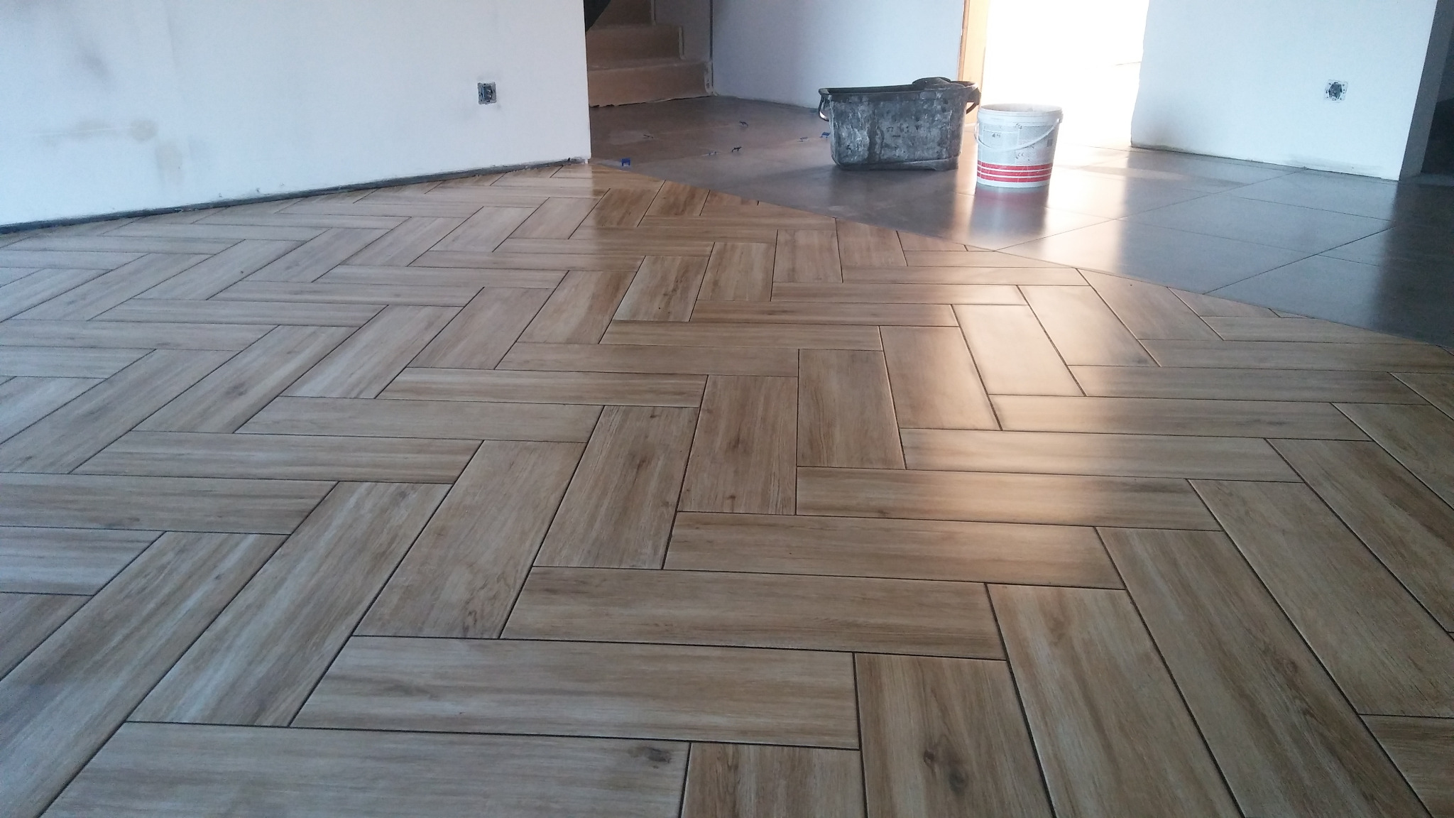 Ułożona pod kątem, drewnopodobna podłoga z płytek, kontrastująca z szarą, gładką powierzchnią. Widoczne wiaderka z zaprawą i fugą oraz fragment ściany z gniazdkiem elektrycznym.