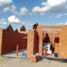 Wznoszenie ścian budynku z czerwonej cegły ceramicznej na placu budowy z widocznym pracownikiem w czarnej koszulce i czerwonej czapce, w tle budynki mieszkalne i błękitne niebo z chmurami.