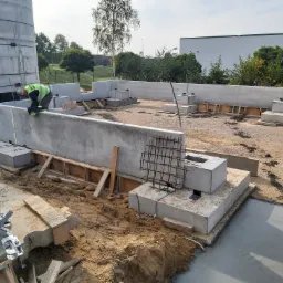 Wylewanie fundamentów pod budynek przemysłowy, widoczny pracownik w kamizelce odblaskowej, szalunki, zbrojenia i betonowe elementy konstrukcyjne, w tle budynek magazynowy.