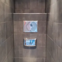 Wąska łazienka wykończona ciemnoszarą, matową glazurą z podtynkową spłuczką i wiszącą toaletą z widocznymi zabrudzeniami na ceramice.