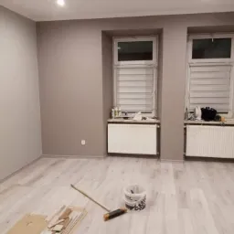 Świeżo pomalowane na szaro wnętrze pokoju z dwoma oknami z żaluzjami, grzejnikami pod oknami, jasną podłogą, widoczne wiadro z farbą oraz pędzle i kartony na podłodze.